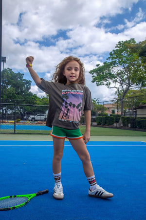 Girl in grey Palm Beach Playground graphic tee and green shorts on a blue tennis court, fist raised – Little Baddie girls streetwear