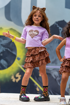 Girl in purple Untamed Heart cropped tee, leopard ruffle skirt, leopard socks and cat ears with face paint in front of a graffiti wall – Little Baddie Halloween